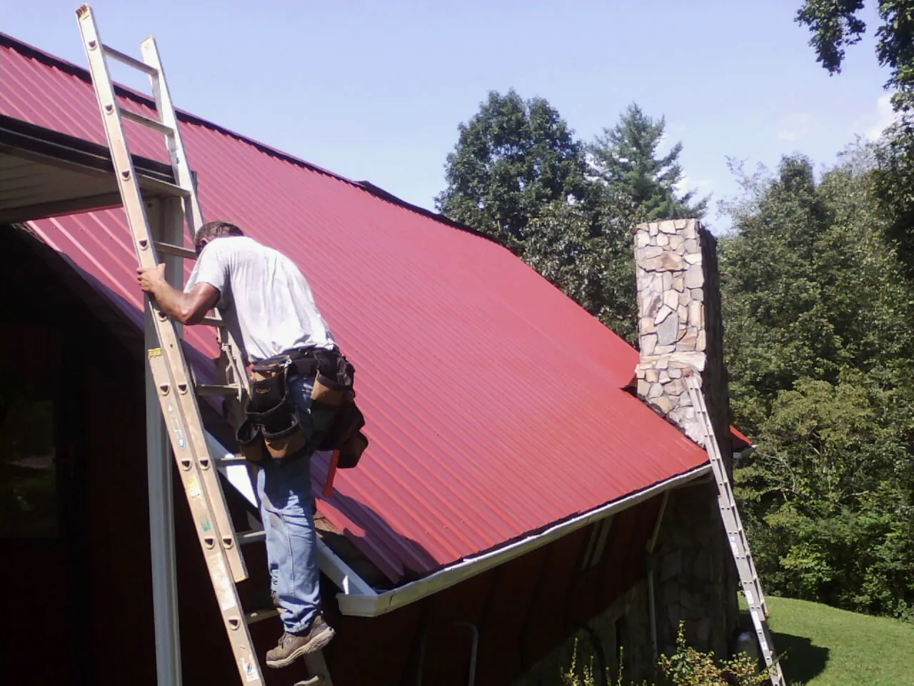 Roofer on a ladder beside a red metal roof and stone chimney — Soffit & Fascia Repair by Roofing Brothers in Esopus